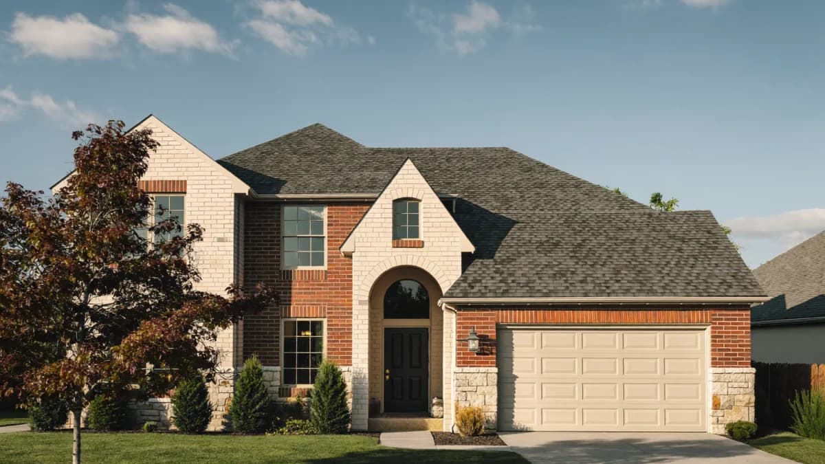 Two-story red brick and cream stone single-family rental home with an arched entry, a two-car garage and a young ornamental tree on a quiet North Texas suburban street in warm afternoon light, the kind of Dallas-Fort Worth house the average rent number is usually wrong about